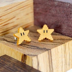 May include: A pair of gold star-shaped earrings with black eyes. The earrings are made of wood and are displayed on a wooden surface.