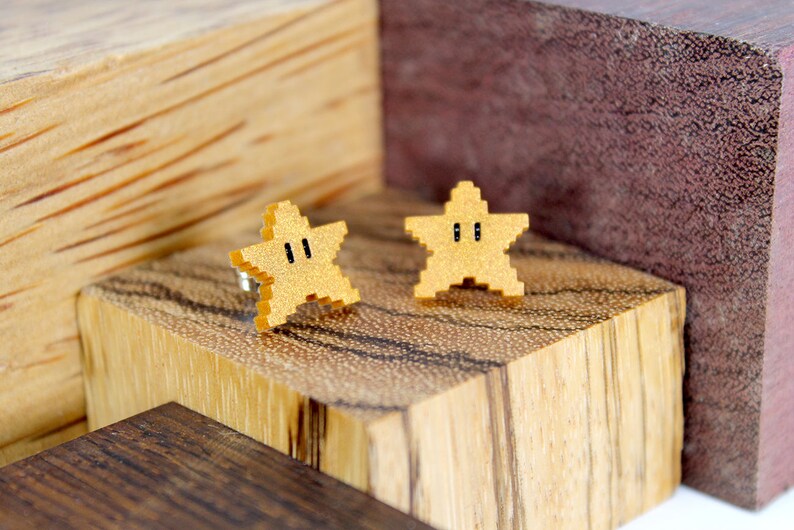 May include: A pair of gold star-shaped earrings with black eyes. The earrings are made of wood and are displayed on a wooden surface.