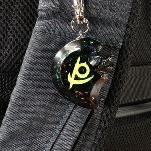 May include: A black and green keychain with a stylized logo. The keychain is attached to a silver chain and is hanging from a black backpack.
