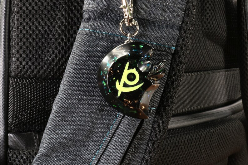 May include: A black and green keychain with a stylized logo. The keychain is attached to a silver chain and is hanging from a black backpack.