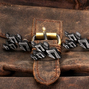 May include: A brown leather bag with a metal buckle and a silver metal design on the front. The design is a series of abstract shapes.