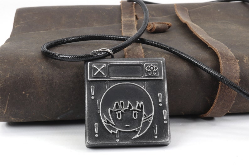May include: A black and silver metal necklace with a square pendant featuring a stylized face with exclamation points around it. The pendant also has a cross and a circle with a dot in the center.