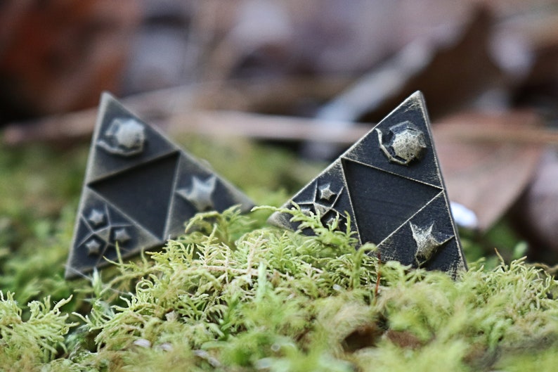 May include: Two black triangular pendants with gold accents and a star design. The pendants are sitting on a bed of green moss.