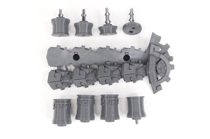 May include: A set of gray 3D printed plastic pieces for a model building kit. The pieces include a long, segmented track, a gear, and several towers.