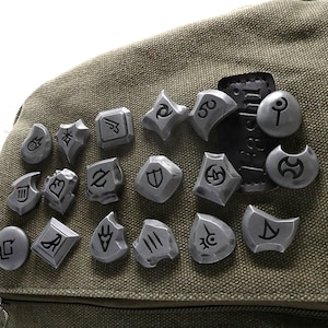 May include: A collection of 18 silver metal tokens with various symbols and designs, arranged on a green canvas bag.
