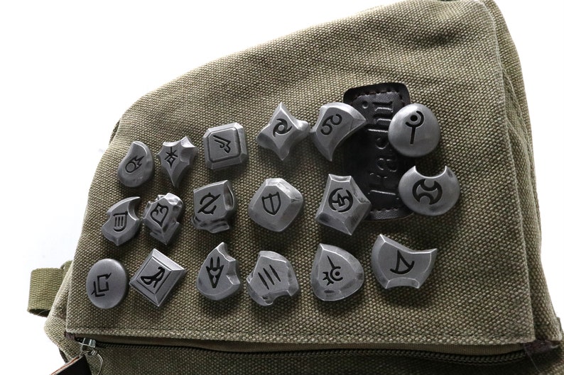 May include: A collection of 18 silver metal tokens with various symbols and designs, arranged on a green canvas bag.