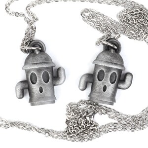 May include: Two silver necklaces with matching pendants. Each pendant is a small, cartoon-like figure with a round head, two eyes, and a surprised expression. The figure is wearing a hat and has two small arms.