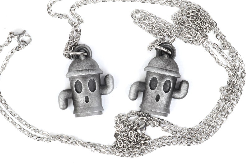 May include: Two silver necklaces with matching pendants. Each pendant is a small, cartoon-like figure with a round head, two eyes, and a surprised expression. The figure is wearing a hat and has two small arms.