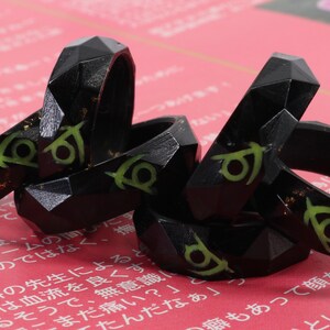 May include: A set of five black resin rings with a green and gold design. The rings are stacked on top of each other and have a geometric shape.