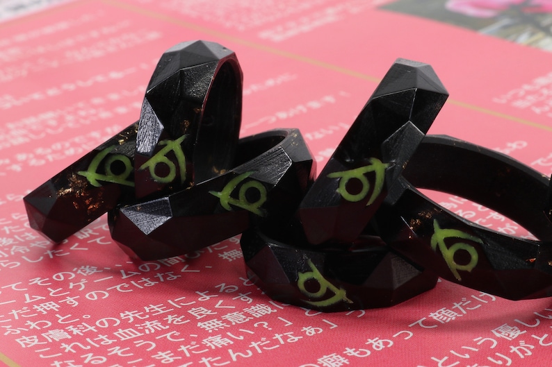 May include: A set of five black resin rings with a green and gold design. The rings are stacked on top of each other and have a geometric shape.