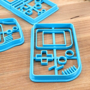 May include: A set of four blue plastic cookie cutters shaped like a handheld video game console. The cutters are on a wooden surface.