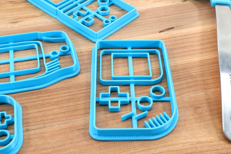 May include: A set of four blue plastic cookie cutters shaped like a handheld video game console. The cutters are on a wooden surface.