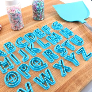 May include: A set of blue plastic alphabet cookie cutters on a wooden surface. The cutters are arranged in alphabetical order.