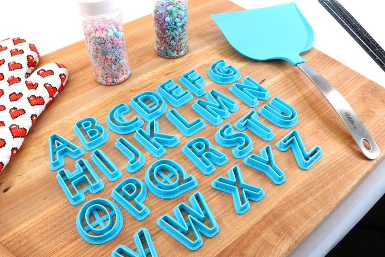 May include: A set of blue plastic alphabet cookie cutters on a wooden surface. The cutters are arranged in alphabetical order.
