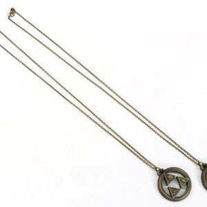 Puede incluir: Dos collares de color dorado con un colgante circular que presenta un dise&ntilde;o triangular estilizado. Los colgantes son id&eacute;nticos y tienen una superficie texturizada.