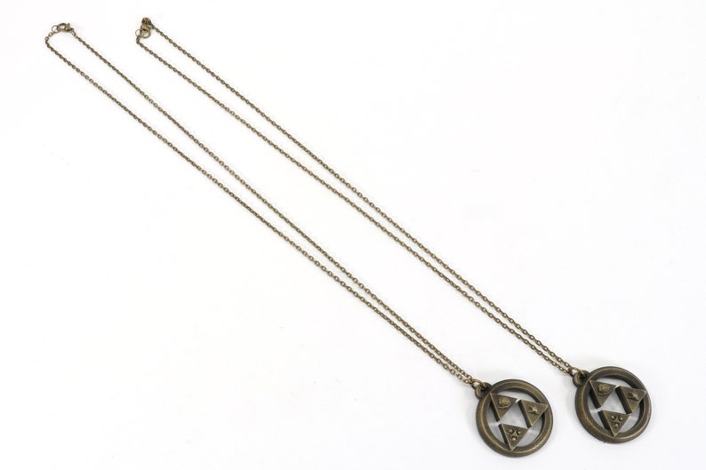 Puede incluir: Dos collares de color dorado con un colgante circular que presenta un dise&ntilde;o triangular estilizado. Los colgantes son id&eacute;nticos y tienen una superficie texturizada.