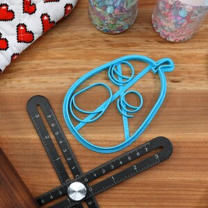 May include: A blue cookie cutter in the shape of a hot air balloon sits on a wooden surface, next to a black multi-angle measuring ruler, a rolling pin, and a blue spatula. A white and red heart-patterned oven mitt is in the background.