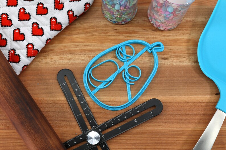 May include: A blue cookie cutter in the shape of a hot air balloon sits on a wooden surface, next to a black multi-angle measuring ruler, a rolling pin, and a blue spatula. A white and red heart-patterned oven mitt is in the background.