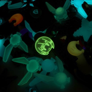 May include: A collection of glow-in-the-dark charms featuring various shapes and designs, including a crescent moon, a cartoon face, and several winged figures. The charms are made of a translucent material that glows brightly in the dark.