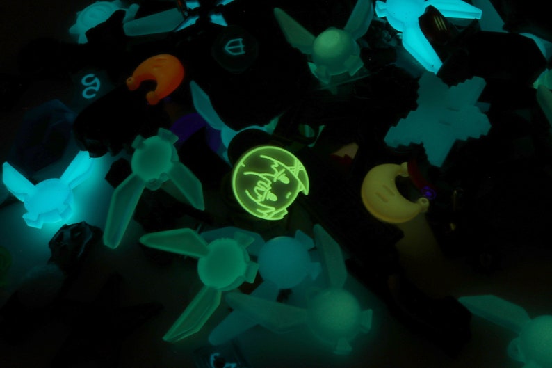 May include: A collection of glow-in-the-dark charms featuring various shapes and designs, including a crescent moon, a cartoon face, and several winged figures. The charms are made of a translucent material that glows brightly in the dark.