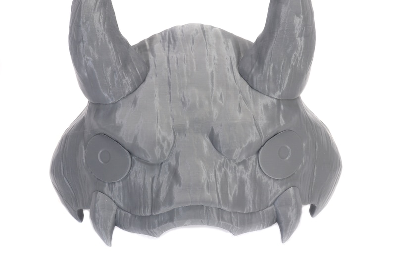 May include: A gray 3D printed mask with horns and two circular eye holes. The mask has a textured surface that resembles wood.