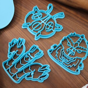May include: Three blue plastic cookie cutters on a wooden surface. The cutters are shaped like a monster, a hand, and a circular design.