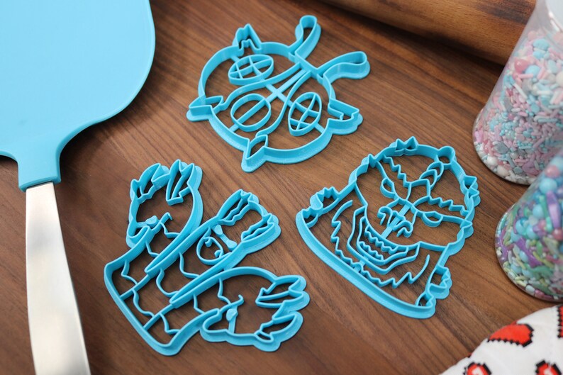 May include: Three blue plastic cookie cutters on a wooden surface. The cutters are shaped like a monster, a hand, and a circular design.