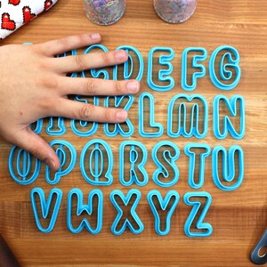May include: A set of blue plastic alphabet cookie cutters on a wooden surface. The cutters are arranged in alphabetical order.