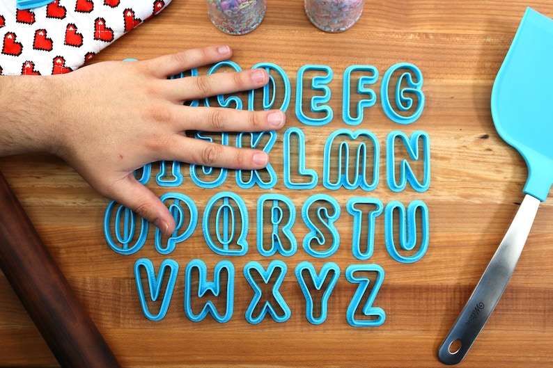 May include: A set of blue plastic alphabet cookie cutters on a wooden surface. The cutters are arranged in alphabetical order.