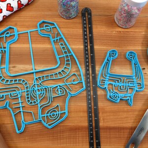 Peut inclure: Deux emportes-pi&egrave;ces en plastique bleu en forme de personnage de jeu vid&eacute;o, un grand et un petit, sur une surface en bois avec une r&egrave;gle, un rouleau &agrave; p&acirc;tisserie et une spatule bleue.
