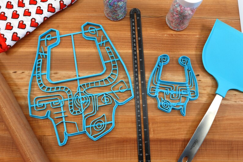 Peut inclure: Deux emportes-pi&egrave;ces en plastique bleu en forme de personnage de jeu vid&eacute;o, un grand et un petit, sur une surface en bois avec une r&egrave;gle, un rouleau &agrave; p&acirc;tisserie et une spatule bleue.