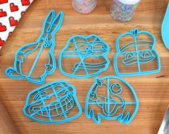 May include: Five blue plastic cookie cutters shaped like internet memes. The cutters are on a wooden surface. The cutters are in the shape of Pepe the Frog, Doge, the 'So you're telling me' meme, the 'Bad Luck Brian' meme, and the 'One Does Not Simply' meme.