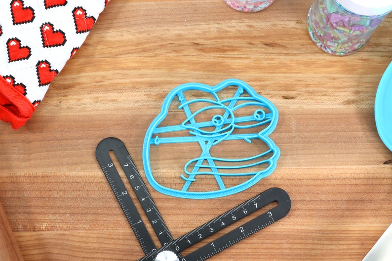 Puede incluir: Un cortador de galletas de pl&aacute;stico azul con forma de rana de dibujos animados con una expresi&oacute;n de sorpresa. El cortador tiene un dise&ntilde;o detallado con l&iacute;neas y c&iacute;rculos.