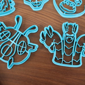 May include: A set of five blue plastic cookie cutters shaped like cartoon monsters. The cutters are on a wooden surface.