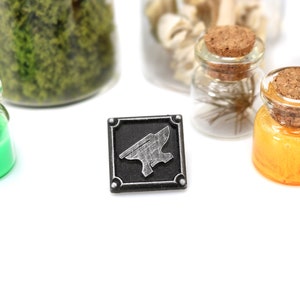 May include: A black and silver metal pin with an anvil design. The pin is on a white surface with several small glass jars with cork tops.
