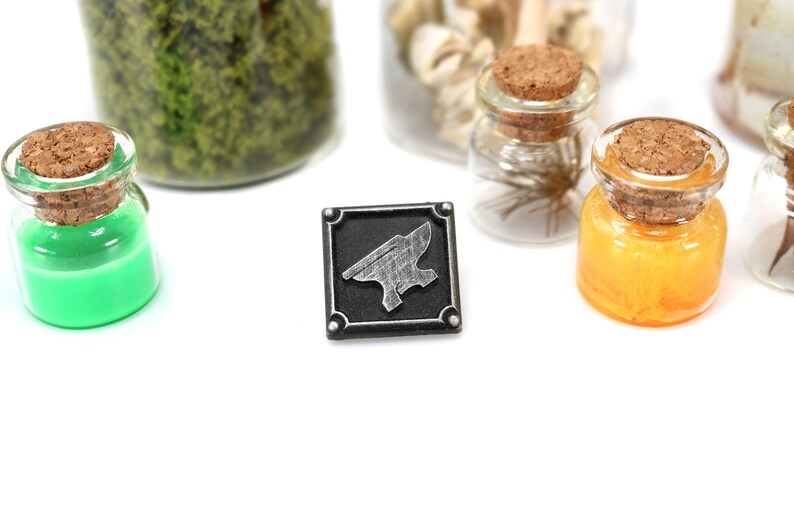 May include: A black and silver metal pin with an anvil design. The pin is on a white surface with several small glass jars with cork tops.