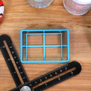 May include: A blue plastic cookie cutter shaped like a grid with four squares. The cutter is on a wooden surface.