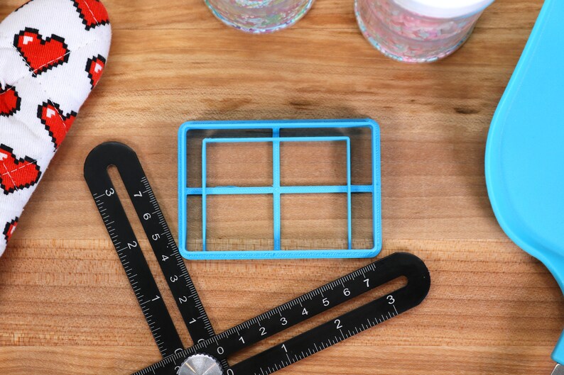 May include: A blue plastic cookie cutter shaped like a grid with four squares. The cutter is on a wooden surface.