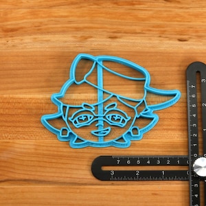 May include: Blue plastic cookie cutter shaped like a cartoon character wearing a hat. The character has large eyes, a small mouth, and a nose.