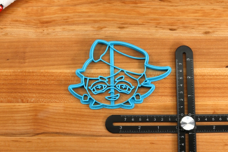 May include: Blue plastic cookie cutter shaped like a cartoon character wearing a hat. The character has large eyes, a small mouth, and a nose.