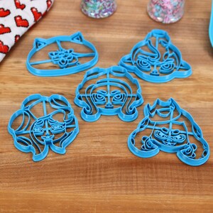 May include: Five blue plastic cookie cutters shaped like cartoon characters with different facial expressions. The cutters are on a wooden surface.