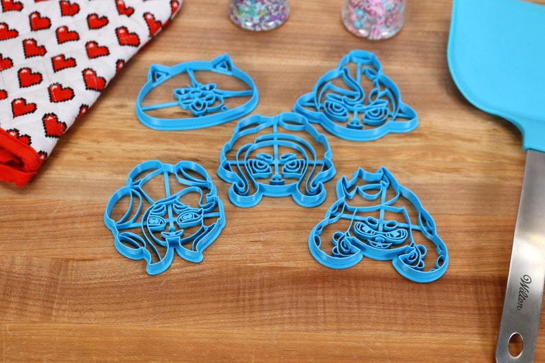 May include: Five blue plastic cookie cutters shaped like cartoon characters with different facial expressions. The cutters are on a wooden surface.