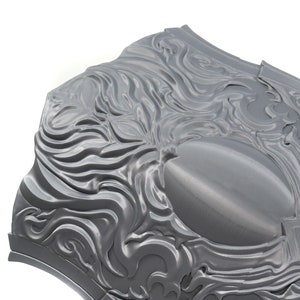 May include: A gray metal shield with a decorative design of swirling, flowing lines. The shield has a smooth, slightly shiny surface.