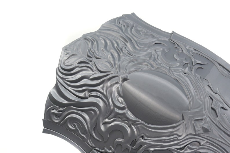 May include: A gray metal shield with a decorative design of swirling, flowing lines. The shield has a smooth, slightly shiny surface.