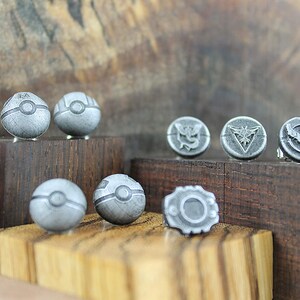 May include: A set of eight silver-colored metal pins featuring Pokemon designs. The pins are displayed on wooden blocks. The designs include Poke Balls and the logos of the three legendary birds, Articuno, Zapdos, and Moltres.