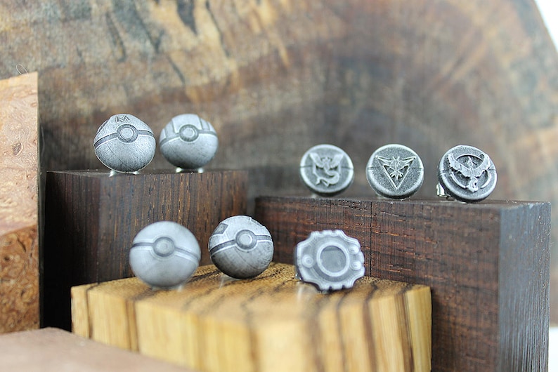 May include: A set of eight silver-colored metal pins featuring Pokemon designs. The pins are displayed on wooden blocks. The designs include Poke Balls and the logos of the three legendary birds, Articuno, Zapdos, and Moltres.