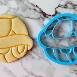 May include: A blue plastic cookie cutter shaped like a turtle shell, next to a yellow cookie in the shape of a turtle shell.