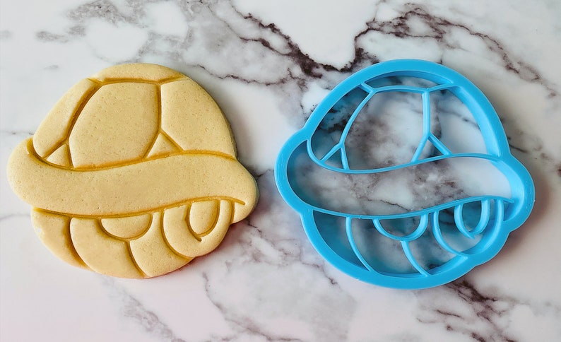 May include: A blue plastic cookie cutter shaped like a turtle shell, next to a yellow cookie in the shape of a turtle shell.