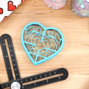 May include: A blue plastic heart-shaped cookie cutter with a decorative design. The cutter is on a wooden surface with other baking supplies.