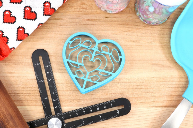 May include: A blue plastic heart-shaped cookie cutter with a decorative design. The cutter is on a wooden surface with other baking supplies.